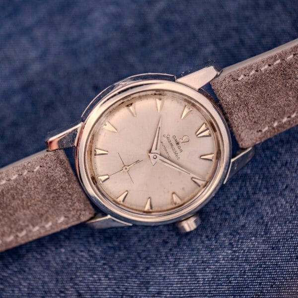 The Omega Automatic Seamaster 2846-2848 features a stainless steel case and sunburst dial, capturing timeless elegance. Displayed on a suede strap against blue fabric, this vintage watch epitomizes classic style.
