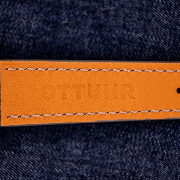 A tan leather strap with OTTUHR embossed, set on dark blue denim, pairs perfectly with the Omega Automatic Seamaster 2846-2848 Stainless Steel featuring a striking Sunburst Dial.