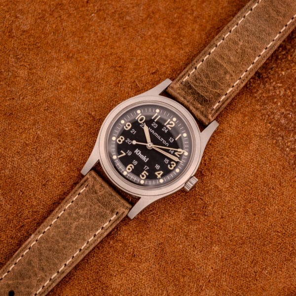 The Hamilton Khaki Ref. 9415A Black Dial Manual Wind Stainless Steel watch with a brown leather strap is displayed on a textured brown surface, highlighting its classic design and manual winding function.