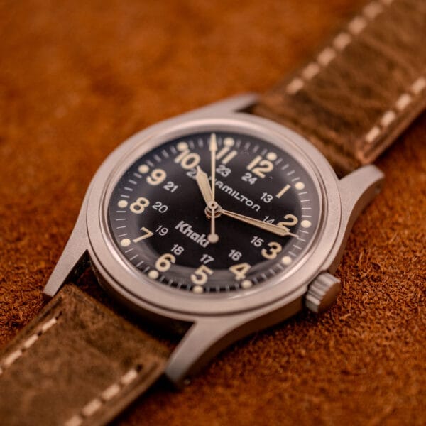 A Hamilton Khaki Ref. 9415A Black Dial Manual Wind Stainless Steel watch with a brown leather strap rests on a textured brown surface.