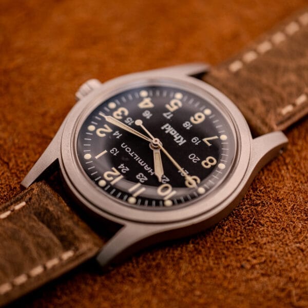 The Hamilton Khaki Ref. 9415A Black Dial Manual Wind Stainless Steel watch with a brown leather strap rests on a textured brown surface, highlighting its classic style.