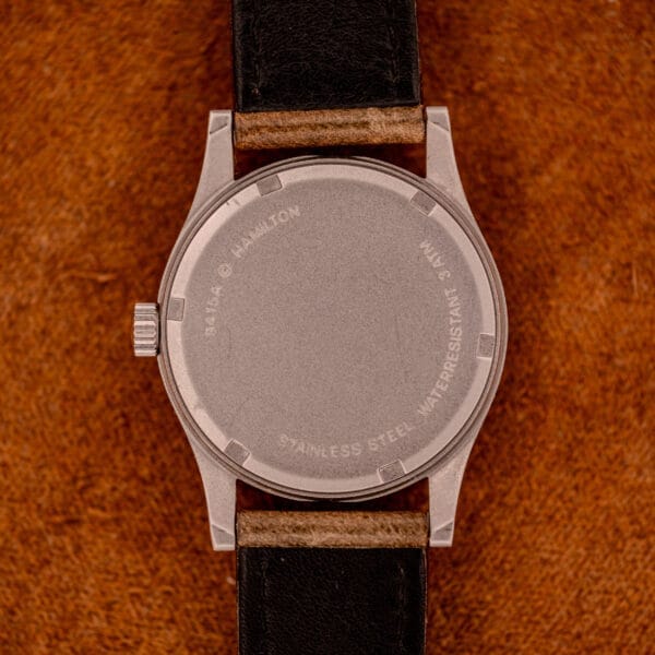 The back of a Hamilton Khaki Ref. 9415A Black Dial Manual Wind Stainless Steel wristwatch with a black leather strap rests on a brown surface, highlighting its classic design.