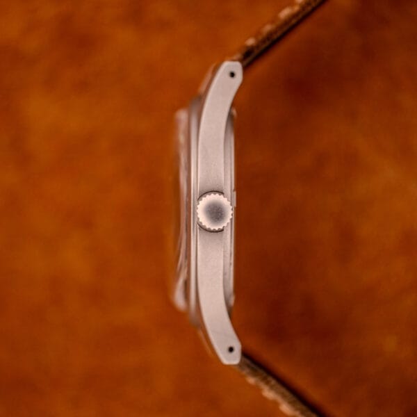 Side view of the Hamilton Khaki Ref. 9415A Black Dial Manual Wind Stainless Steel watch with a brown leather strap, featuring its silver crown against a blurred brown background.