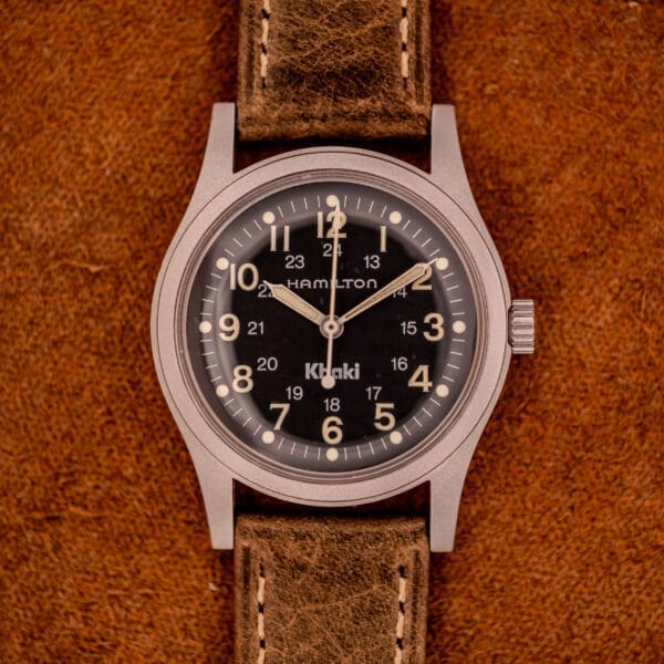 A Hamilton Khaki Ref. 9415A Black Dial Manual Wind Stainless Steel wristwatch with a brown leather strap rests on a brown suede surface.