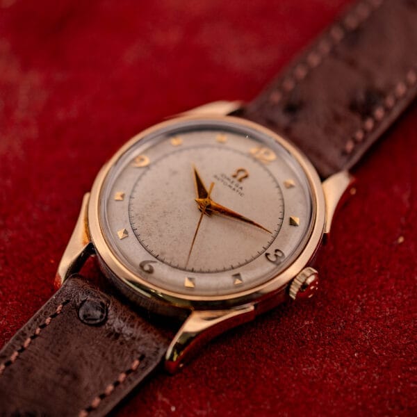 Omega Automatic Bumper Ref. 2582 14k Gold Capped Cal. 351 wristwatch with a brown leather strap, displayed on a red textured surface.