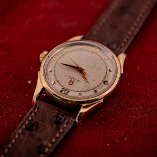 Omega Automatic Bumper Ref. 2582 wristwatch with 14k gold capped case and Cal. 351 movement, featuring a brown leather strap, displayed on a red textured surface.