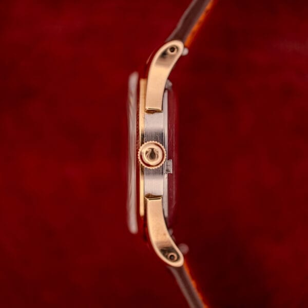 Side view of an Omega Automatic Bumper Ref. 2582 14k Gold Capped Cal. 351 wristwatch with a brown strap against a red textured background.