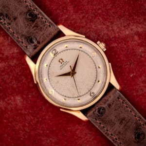 The Omega Automatic Bumper Ref. 2582 features a 14k gold capped case and Cal. 351 movement, paired with a brown leather strap, displayed on a red textured background.