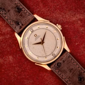 The Omega Automatic Bumper Ref. 2582 features a 14k gold capped case and Cal. 351 movement, paired with a brown leather strap, displayed on a red textured background.
