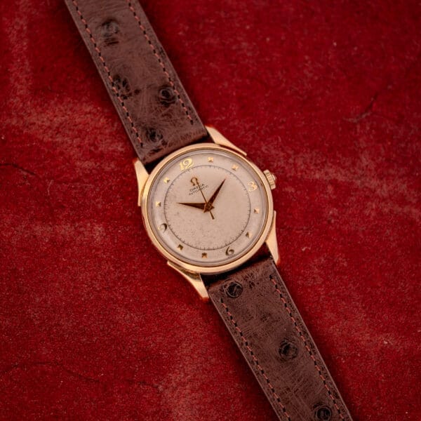An Omega Automatic Bumper Ref. 2582 with a brown leather strap and 14k gold capped case (Cal. 351), shown on a red textured surface.