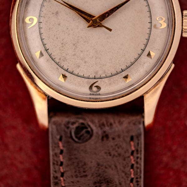 Close-up of an Omega Automatic Bumper Ref. 2582 14k Gold Capped watch with Cal. 351 movement, worn white dial, and brown leather strap on a red background.