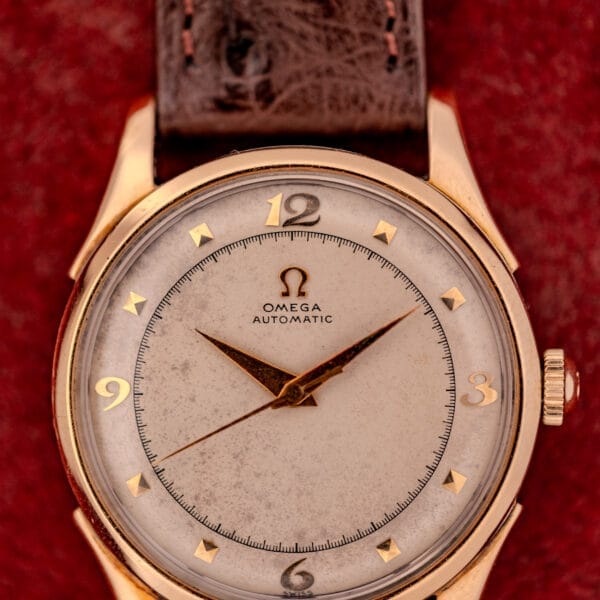 The Omega Automatic Bumper Ref. 2582 features a 14k gold capped case, brown leather strap, and Cal. 351 movement, beautifully displayed against a striking red background.