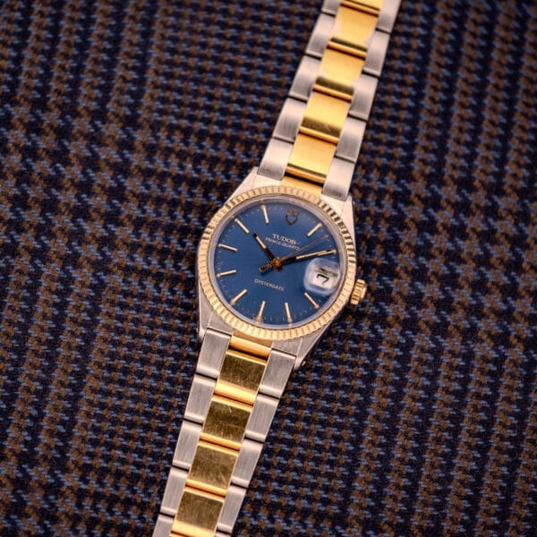 A Tudor Prince-Quartz Oysterdate Ref. 9150 two-tone wristwatch with a blue dial rests on brown and blue houndstooth fabric.