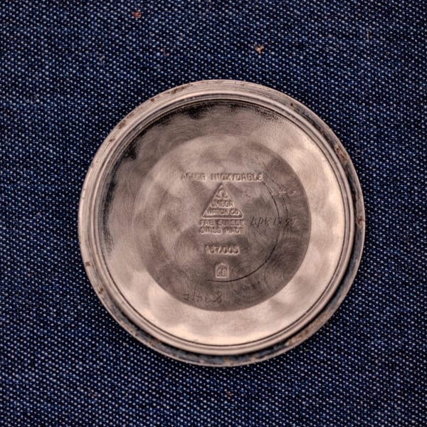 The round metallic back of the Omega Constellation Pie-Pan Dial Ref. 167.005 14k Gold Capped, engraved with text, rests on a blue textured fabric background.