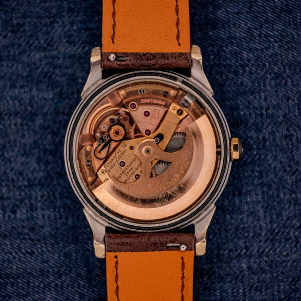 An Omega Constellation Pie-Pan Dial Ref. 167.005 wristwatch with visible mechanical movement and a brown leather strap rests on blue denim fabric.