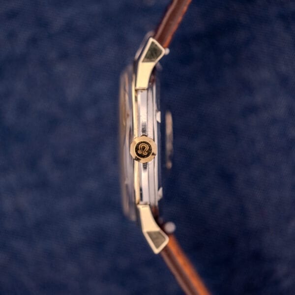 Side view of an Omega Constellation Pie-Pan Dial Ref. 167.005 14k Gold Capped wristwatch with a brown leather strap and Omega logo on the crown, set against a blue background.