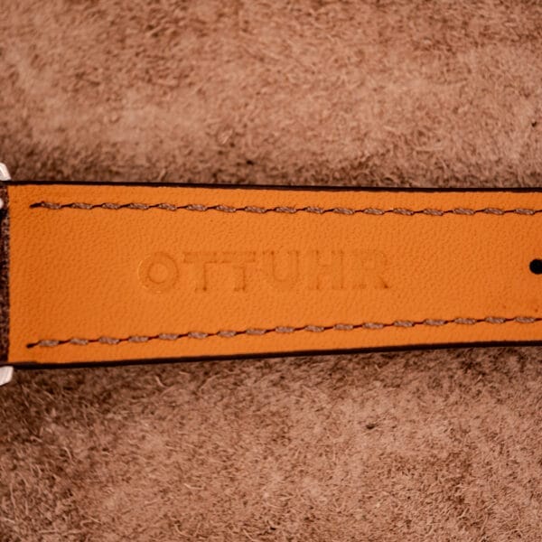 Close-up of a brown leather strap embossed with "OTTUHR" on a textured surface, next to a Tudor Oysterdate Ref. 90120 Stainless Steel Sunburst Dial case.