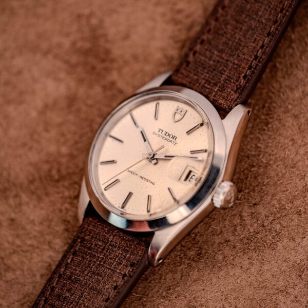 A Tudor Oysterdate Ref. 90120 Stainless Steel Sunburst Dial wristwatch with a brown strap rests on a brown textured surface, emphasizing its timeless design and sunburst dial elegance.