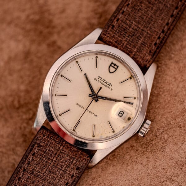 Tudor Oysterdate Ref. 90120 in stainless steel, featuring a beige sunburst dial, date at 3 o’clock, and a brown textured strap.