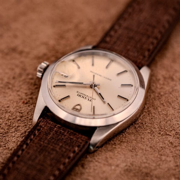 A Tudor Oysterdate Ref. 90120 Stainless Steel Sunburst Dial wristwatch with a brown strap rests on a textured brown surface, highlighting its classic case design.