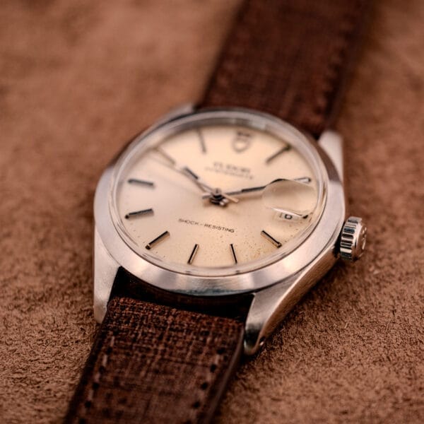 A Tudor Oysterdate Ref. 90120 Stainless Steel Sunburst Dial wristwatch with a brown leather strap rests on suede, its date window shown magnified.