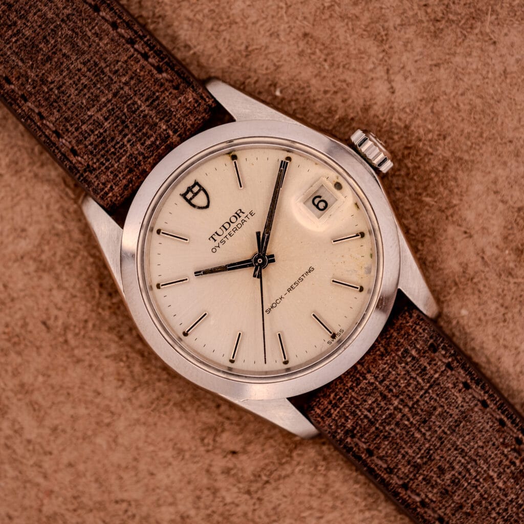 A Tudor Oysterdate Ref. 90120 Stainless Steel watch with a sunburst dial, date window, and brown textured strap displayed on a tan background.