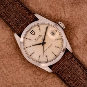 A Tudor Oysterdate Ref. 90120 Stainless Steel watch with a sunburst dial, date window, and brown textured strap displayed on a tan background.