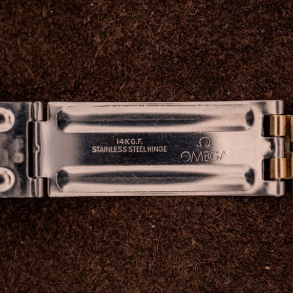 Close-up of an Omega Constellation Chronometer Ref. 168.045 Day/Date Full Set clasp, engraved with “14K G.F. Stainless Steel Hinge OMEGA 168.045” on a brown background, emphasizing its fine chronometer craftsmanship.