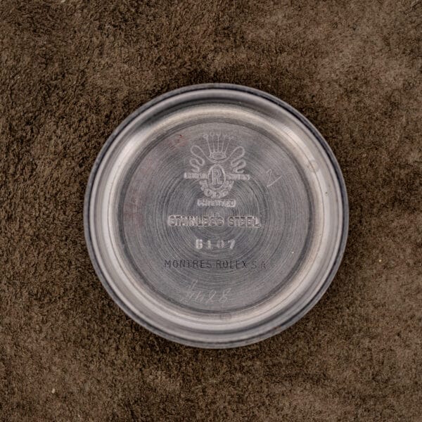 A round stainless steel case back with Rolex engravings from a Rolex Oyster Perpetual "Big Bubble Back" Ref. 6107 Black Dial is displayed on a brown textured surface.