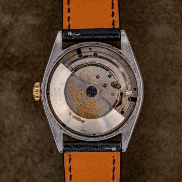 Close-up of a Rolex Oyster Perpetual "Big Bubble Back" Ref. 6107 Black Dial showing its vintage movement, exposed gears and rotor, paired with a tan leather strap on a brown surface.