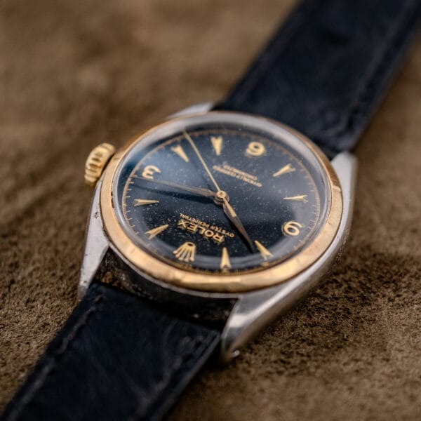 Close-up of the Rolex Oyster Perpetual "Big Bubble Back" Ref. 6107 Black Dial, featuring gold accents and a black strap, displayed on a brown surface.