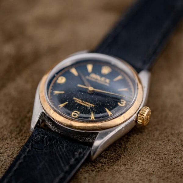 Close-up of the Rolex Oyster Perpetual "Big Bubble Back" Ref. 6107 Black Dial, shown with a gold bezel and black leather strap on a textured brown surface.