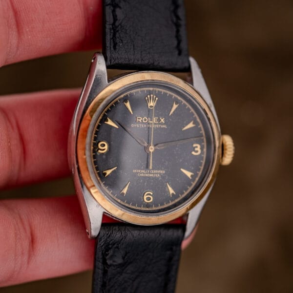 A hand holds a Rolex Oyster Perpetual "Big Bubble Back" Ref. 6107 Black Dial watch with a black leather strap and gold-tone details.