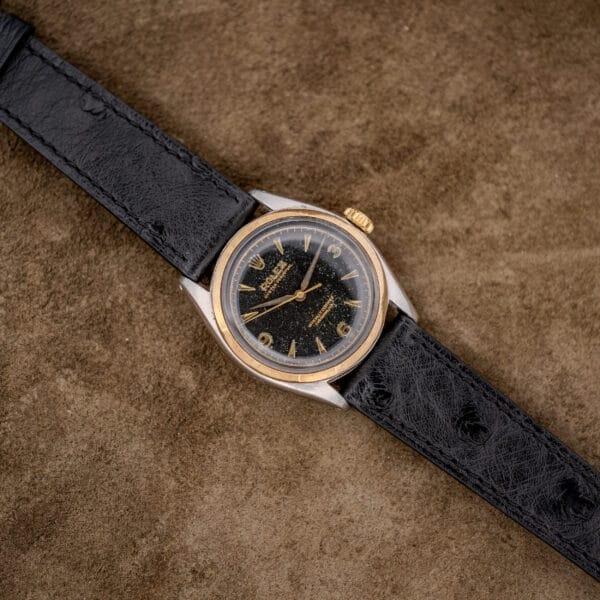 Rolex Oyster Perpetual "Big Bubble Back" Ref. 6107 with a black dial and leather strap, displayed on a brown textured surface.