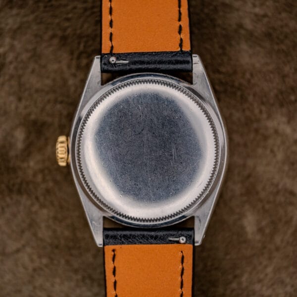 A close-up of the back of a Rolex Oyster Perpetual "Big Bubble Back" Ref. 6107 Black Dial with a tan and black leather strap, resting on a brown textured surface.