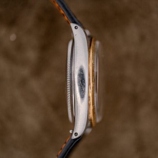 Side view of a worn Rolex Oyster Perpetual "Big Bubble Back" Ref. 6107 Black Dial with a scratched metallic case and leather strap against a brown background.