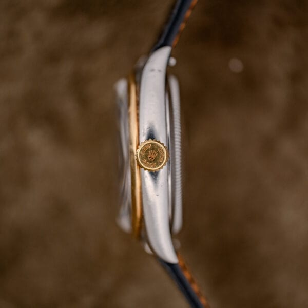 Side view of a Rolex Oyster Perpetual "Big Bubble Back" Ref. 6107 Black Dial, highlighting its gold crown with the Rolex logo set against a blurred brown background.