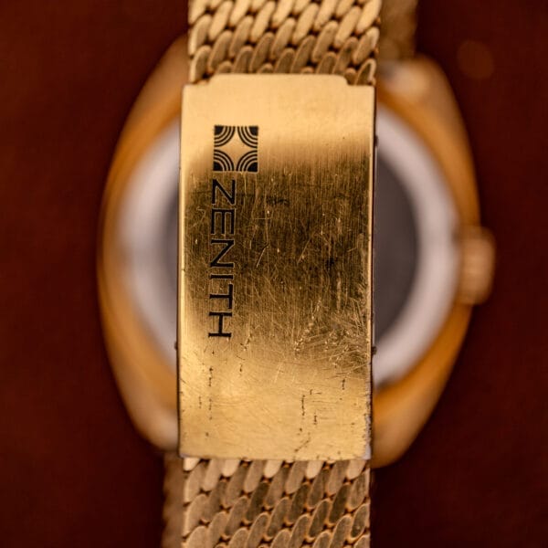 Close-up of a scratched gold Movado / Zenith Electronic Tuning Fork Cal ESA 1962 watch clasp with textured metal band and faint watch face, echoing early tuning fork technology; includes original box and papers.