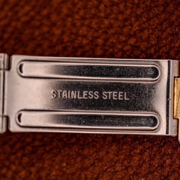 Close-up of a metal clasp engraved "STAINLESS STEEL" against a brown backdrop—reflecting the refined craftsmanship found in the Movado / Zenith Electronic Tuning Fork Cal ESA 1962 Box & Papers.