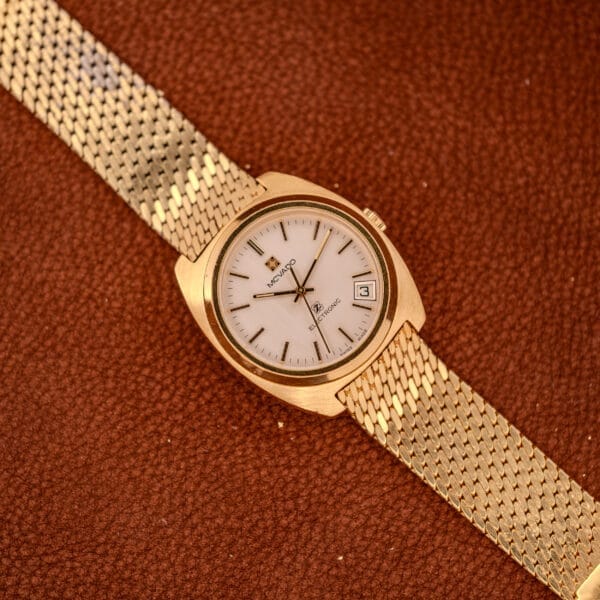 Movado / Zenith Electronic Tuning Fork Cal ESA 1962 wristwatch with mesh band on a brown textured surface, showcasing date and time—timeless style with original box & papers.