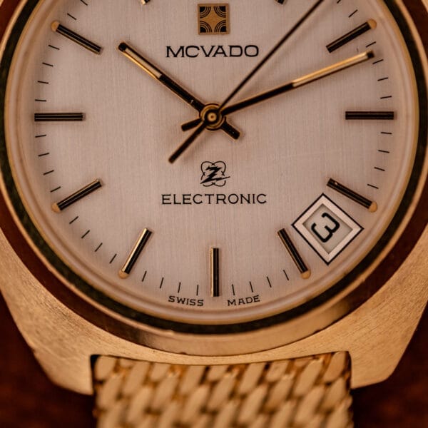 Close-up of a gold Movado / Zenith Electronic Tuning Fork Cal ESA 1962 wristwatch, showing the time as 1:19 and the date as 3, sold with box and papers.