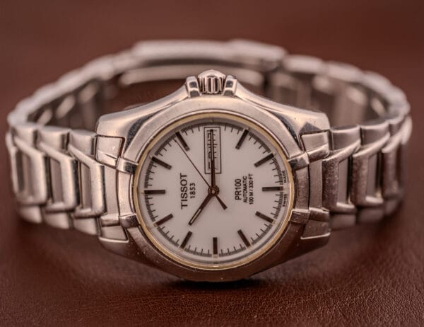 A Tissot PR100 Automatic Day/Date P 764 in stainless steel with a white dial and metal band rests on a brown surface.