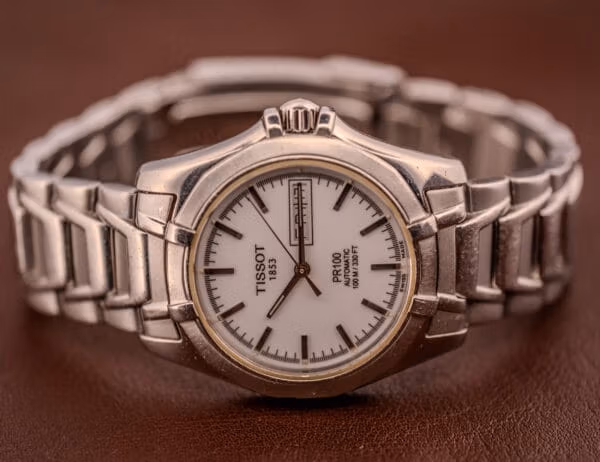 Tissot PR100 Automatic Day/Date Ref. P 764 resting on brown leather with the signed steel bracelet curled to either side of the case.