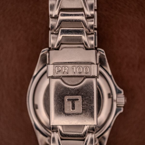 Close-up of a Tissot PR100 Automatic Day/Date P 764 stainless steel clasp, engraved with "PR 100" and a large "T", set against a brown background.