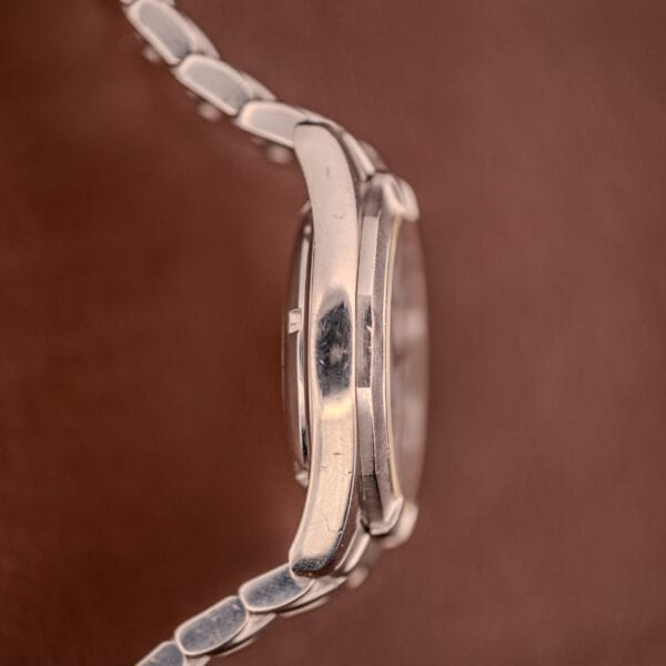 Side view of the Tissot PR100 Automatic Day/Date P 764 in stainless steel, featuring an automatic day/date display, set against a brown textured background.