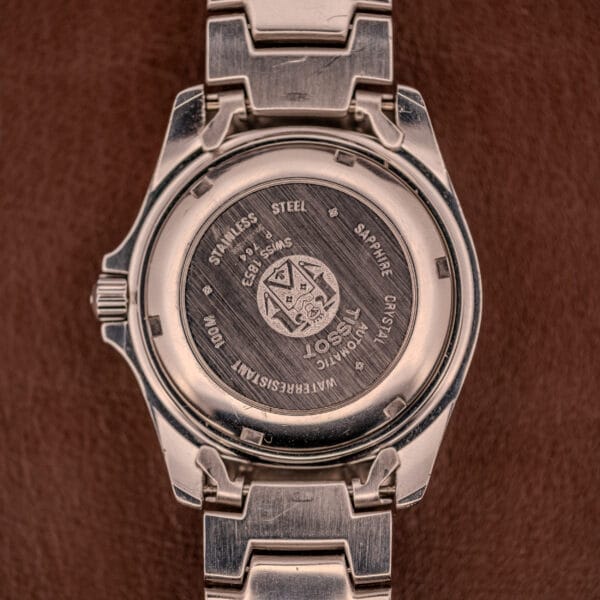 The back of a Tissot PR100 Automatic Day/Date P 764 stainless steel wristwatch features detailed engravings, displayed against a brown leather background.
