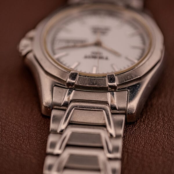 Close-up of the Tissot PR100 Automatic Day/Date P 764 wristwatch in stainless steel with a silver dial and band, resting on a brown surface.