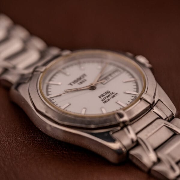 A close-up of the Tissot PR100 Automatic Day/Date P 764 wristwatch in stainless steel with a white face and metal band, displayed on a brown surface.