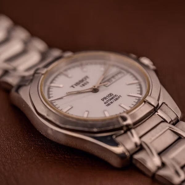 Front-leaning macro of the Tissot PR100 dial highlighting the applied steel batons, day-date window, and PR100 AUTOMATIC printing.