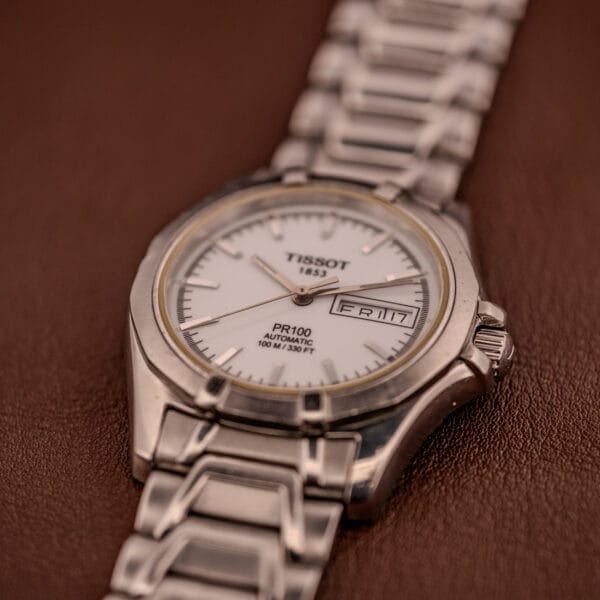 The Tissot PR100 Automatic Day/Date P 764 in stainless steel features a white dial and silver band, shown on a brown surface.